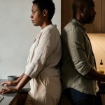 A man and woman stand back to back in a modern kitchen. The woman is African, middle-aged, with short hair. The man is African, middle-aged, with a beard.