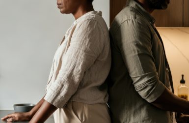 A man and woman stand back to back in a modern kitchen. The woman is African, middle-aged, with short hair. The man is African, middle-aged, with a beard.