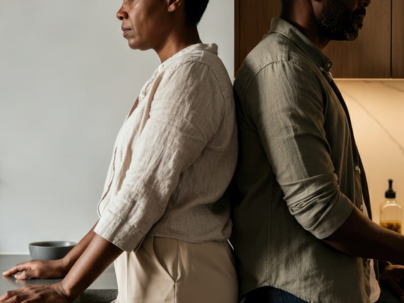 A man and woman stand back to back in a modern kitchen. The woman is African, middle-aged, with short hair. The man is African, middle-aged, with a beard.